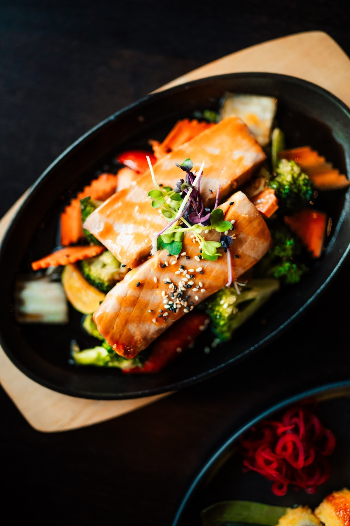 Gebratener Lachs mit Sesam und Sprossen auf gebratenem Gemüse wie Brokkoli, Karotten, Paprika und Aubergine, serviert auf einem schwarzen Teller.